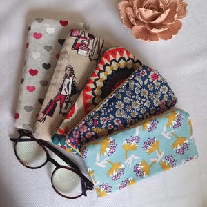 May include: Five rectangular fabric cases with various patterns including red and gray hearts, a person shopping, geometric red and black shapes, blue floral, and light blue with yellow and purple flowers, alongside dark brown tortoise shell glasses.