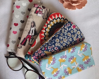 May include: Five rectangular fabric cases with various patterns including red and gray hearts, a person shopping, geometric red and black shapes, blue floral, and light blue with yellow and purple flowers, alongside dark brown tortoise shell glasses.