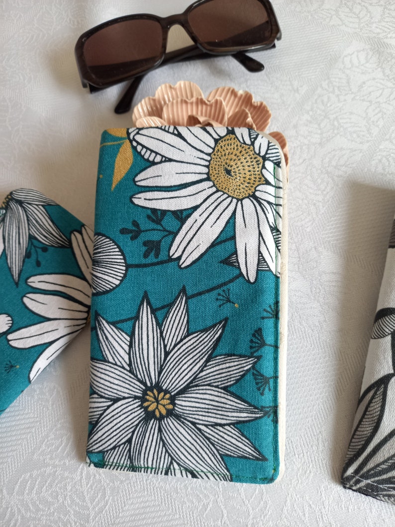 May include: A teal blue fabric glasses case with a white and black floral pattern. The flowers have yellow centers. The case is closed.