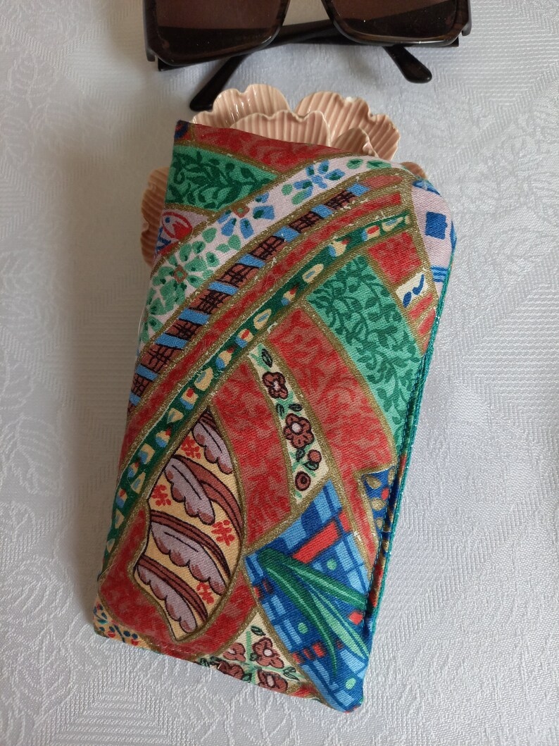 May include: A red, green, blue, and gold patterned fabric eyeglass case. The case is made of a soft, quilted fabric and has a zipper closure.