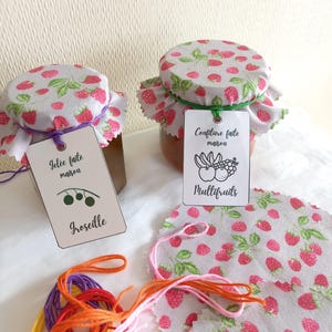 Puede incluir: Dos tarros de vidrio con mermelada y jalea caseras. Los tarros están cubiertos con tapas de tela con un patrón de fresas. Las etiquetas dicen "Confiture faite maison Multifruits" y "Gelée faite maison Groseille".