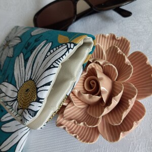 May include: A teal and white floral patterned glasses case with a white satin lining. The case is folded closed and is lying on a white surface with a pink ceramic rose.