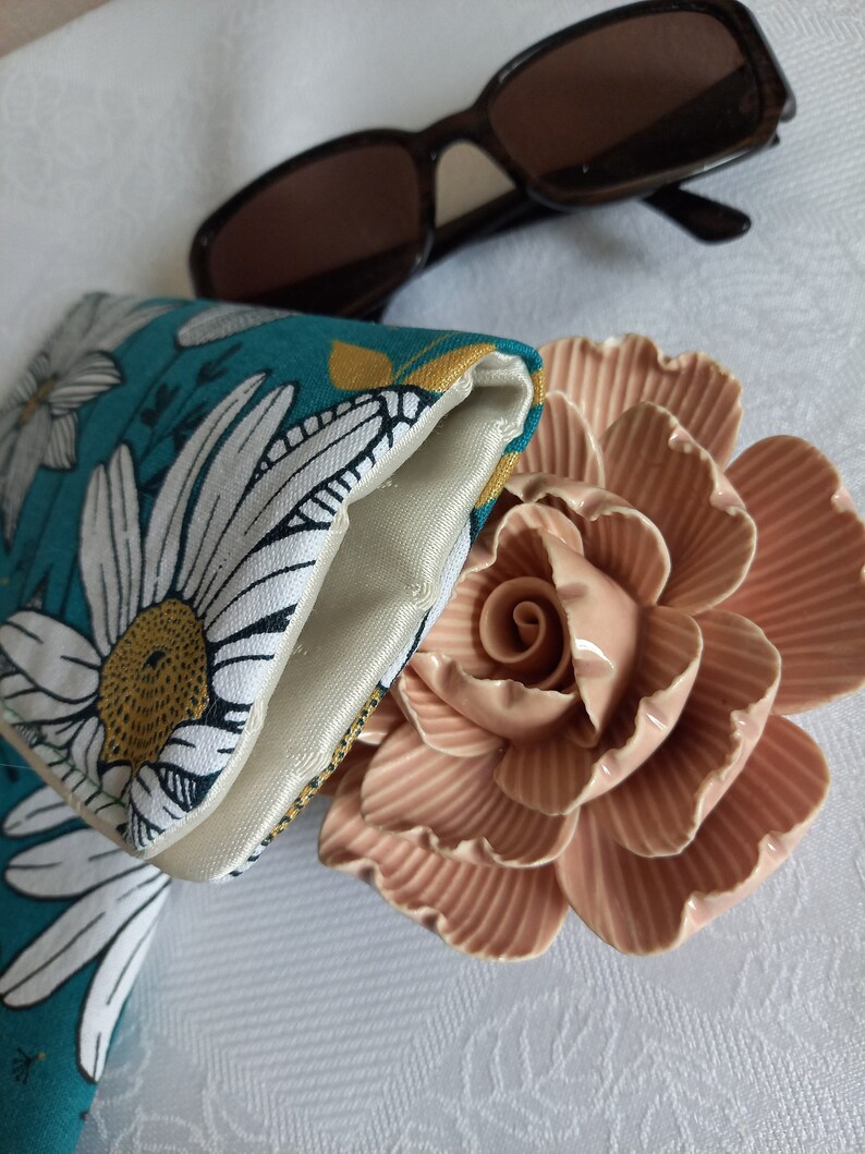May include: A teal and white floral patterned glasses case with a white satin lining. The case is folded closed and is lying on a white surface with a pink ceramic rose.