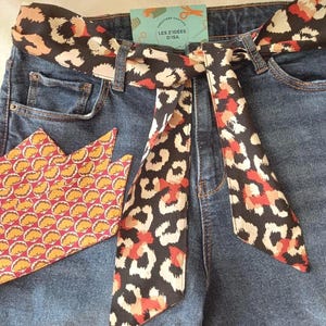 Peut inclure: Un jean en denim bleu foncé avec une ceinture en tissu à motif léopard noir, blanc et orange nouée à la taille. Une pièce de tissu triangulaire séparée avec un motif en éventail jaune, rose et blanc est posée à gauche. Une étiquette indique LES Z'IDÉES D'ISA.