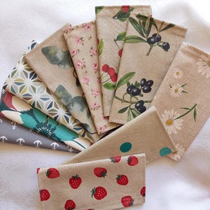 May include: A collection of rectangular fabric napkins arranged in a fan shape. The napkins feature various prints including red strawberries, white daisies, black olives, pink roses, green leaves, geometric patterns, teal flowers, and grey anchors.