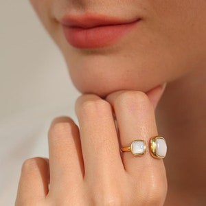 May include: A gold ring with two white square-shaped stones. The ring is open on one side and the stones are set in a bezel setting.