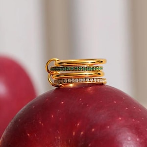 Könnte beinhalten: Ein Stapel von vier goldenen Ringen, jeder mit einem anderen Design, liegt auf einem roten Apfel. Ein Ring hat eine Reihe grüner Edelsteine, ein anderer ist mit kleinen, klaren Steinen besetzt. Die Ringe haben eine glänzende, polierte Oberfläche.