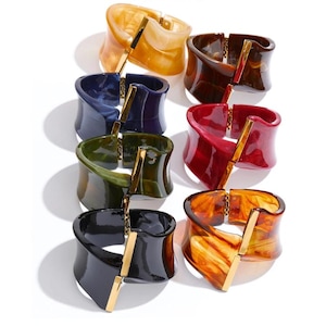 Op de afbeelding: Een verzameling van acht kleurrijke, dikke armbanden. Elke armband heeft een gebogen, brede band in tinten amber, bruin, rood, blauw, groen en zwart. Een goudkleurige metalen staaf en scharnier verbinden de uiteinden van de armband.