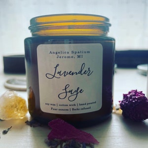 Op de afbeelding: Een kaars in een amberkleurige glazen pot met een wit etiket met de tekst Angelica Spatium Jerome, MI, Lavender Sage, soy wax, cotton wick, hand poured, four ounces, Reiki infused. De kaars staat naast gedroogde lavendel en een kristal.