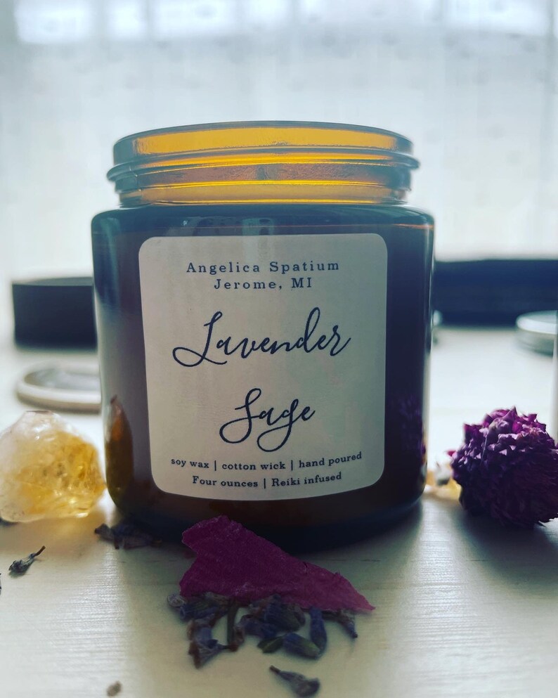 Op de afbeelding: Een kaars in een amberkleurige glazen pot met een wit etiket met de tekst Angelica Spatium Jerome, MI, Lavender Sage, soy wax, cotton wick, hand poured, four ounces, Reiki infused. De kaars staat naast gedroogde lavendel en een kristal.