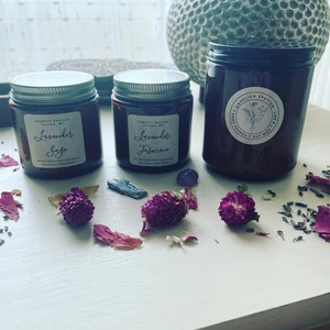 Op de afbeelding: Drie amberkleurige glazen kaarsenhouders met zilveren of zwarte deksels en witte etiketten. Twee kleinere potten zijn gelabeld met Lavender Sage en Lavender Jasmine. De grotere pot heeft een Angelica Spatium logo. Paarse bloemen en kleine kristallen liggen rondom de potten.