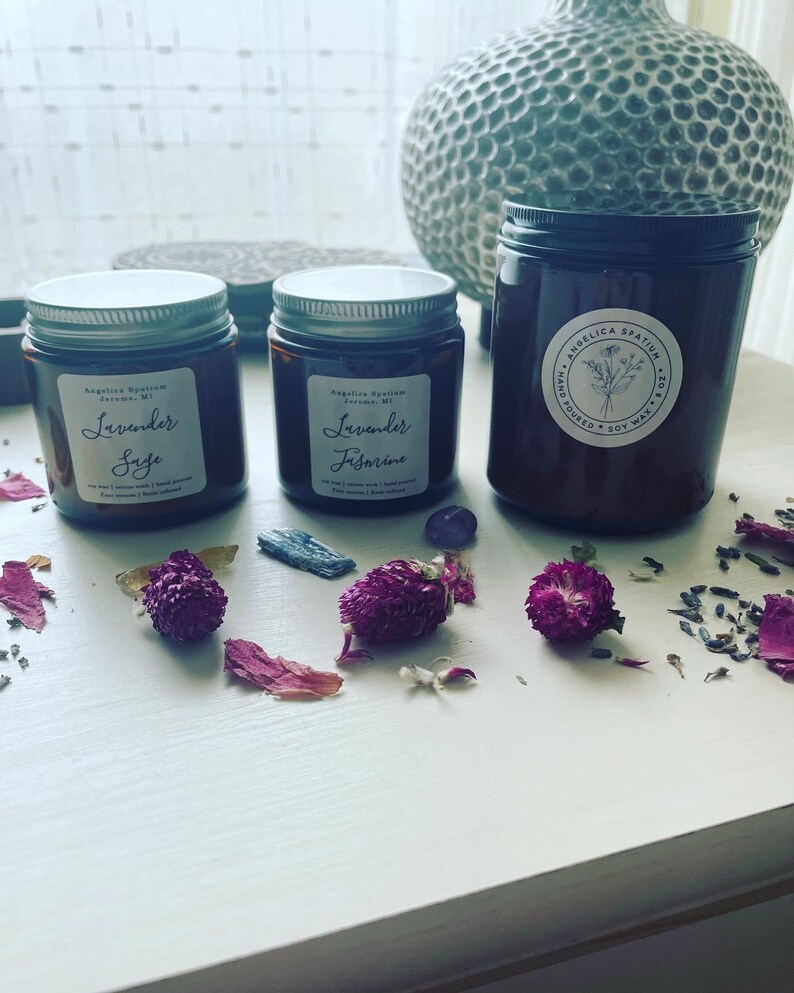 Op de afbeelding: Drie amberkleurige glazen kaarsenhouders met zilveren of zwarte deksels en witte etiketten. Twee kleinere potten zijn gelabeld met Lavender Sage en Lavender Jasmine. De grotere pot heeft een Angelica Spatium logo. Paarse bloemen en kleine kristallen liggen rondom de potten.