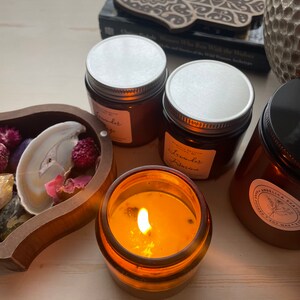 Op de afbeelding: Verschillende amberkleurige glazen kaarsenhouders met zilveren en zwarte deksels, sommige met witte etiketten met de tekst Lavender Sage en Lavender Jasmine. E&eacute;n kaars brandt met een gele vlam. Een houten dienblad bevat gedroogde bloemen en een schijfje agaat.