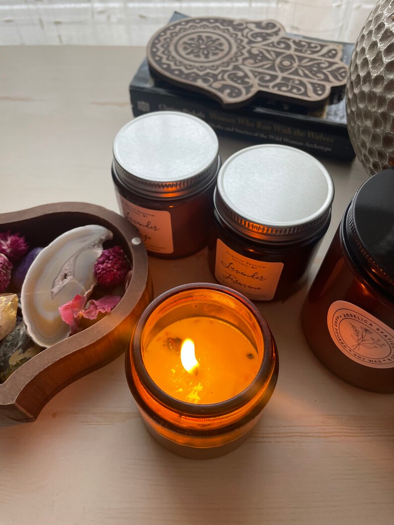 Op de afbeelding: Verschillende amberkleurige glazen kaarsenhouders met zilveren en zwarte deksels, sommige met witte etiketten met de tekst Lavender Sage en Lavender Jasmine. E&eacute;n kaars brandt met een gele vlam. Een houten dienblad bevat gedroogde bloemen en een schijfje agaat.