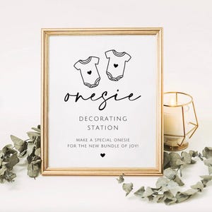 May include: A framed sign with black text and graphics on a white background. The sign features two baby onesie illustrations, the text "onesie DECORATING STATION", and the phrase "MAKE A SPECIAL ONESIE FOR THE NEW BUNDLE OF JOY!" with a small heart icon.