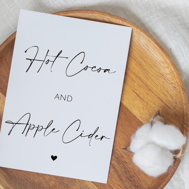 May include: A white card with the words "Hot Cocoa AND Apple Cider" in elegant black script, with a small black heart at the bottom. The card rests on a wooden tray, with a cotton ball on the side.