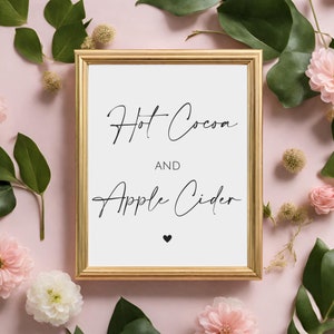 May include: A framed sign with gold trim displays the words "Hot Cocoa AND Apple Cider" in elegant black script, with a small black heart beneath. The sign is surrounded by pink flowers and green leaves on a pink background.