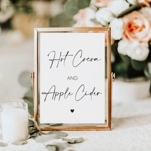 May include: A framed sign with the words "Hot Cocoa AND Apple Cider" in elegant black script. The sign is in a gold frame, with a small black heart at the bottom. A white candle and floral arrangement are in the background.