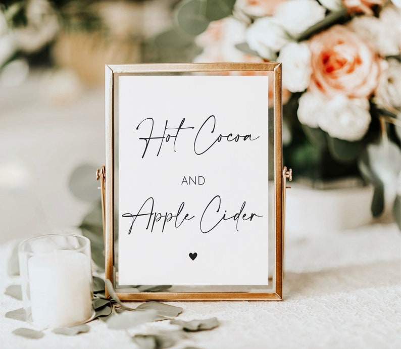 May include: A framed sign with the words "Hot Cocoa AND Apple Cider" in elegant black script. The sign is in a gold frame, with a small black heart at the bottom. A white candle and floral arrangement are in the background.