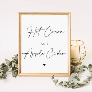 May include: A framed sign with gold trim displays the words "Hot Cocoa AND Apple Cider" in elegant black script. A small black heart is below the text. A lit candle in a gold geometric holder and greenery are in the background.