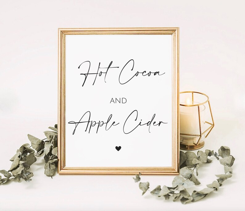 May include: A framed sign with gold trim displays the words "Hot Cocoa AND Apple Cider" in elegant black script. A small black heart is below the text. A lit candle in a gold geometric holder and greenery are in the background.