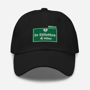 May include: Black baseball cap with a green and white embroidered patch that reads "TRACK 13 In Stilettos 4 Miles" with a broken heart icon.