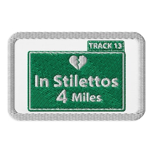 May include: A white embroidered patch with a green road sign that says "In Stilettos 4 Miles" with a broken heart icon above the text. The sign has a small white label that says "Track 13".