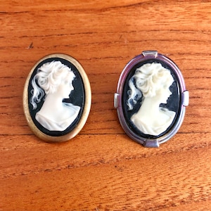 May include: Two oval cameo brooches with a white cameo of a woman's profile set against a black background. One brooch has a gold-toned frame and the other has a silver-toned frame.