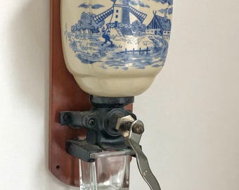 以下が含まれることがあります： 青い白の風車を描いたセラミック製の壺が付いた、ヴィンテージの壁掛け式コーヒーグラインダー。グラインダーは黒い金属製の機構と木製のクランクハンドルが付いています。