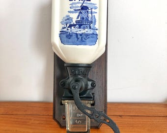 以下が含まれることがあります： 青い白のセラミック製のジャーに「CAFÉ」と風車のデザインが描かれた、ヴィンテージの壁掛け式コーヒーグラインダー。グラインダーは黒い金属製で、木製のベースが付いています。
