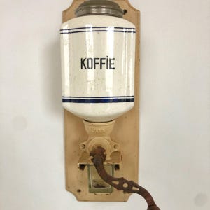 以下が含まれることがあります： 「KOFFIE」と書かれた白い陶器の壺と錆びた金属製のクランクが付いた、ヴィンテージの壁掛け式コーヒーグラインダーです。