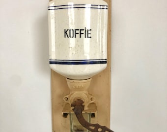 以下が含まれることがあります： 「KOFFIE」と書かれた白い陶器の壺と錆びた金属製のクランクが付いた、ヴィンテージの壁掛け式コーヒーグラインダーです。