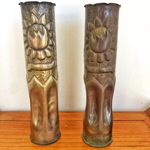 Peut inclure: Deux grands vases cylindriques en laiton avec une texture martelée et un motif de tournesol en relief sur la partie supérieure. Chaque vase présente un bord festonné et une base lisse et effilée, avec une patine métallique dorée.