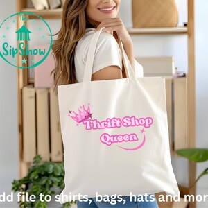 Puede incluir: Una bolsa de tela blanca con una corona rosa y el texto "Thrift Shop Queen" en letras rosas. La bolsa está siendo sostenida por una persona.