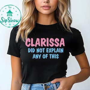 May include: Black t-shirt with the text "Clarissa Did Not Explain Any Of This" in pink and blue. The shirt also features a circular logo with text and stars. The shirt is a casual wear item.
