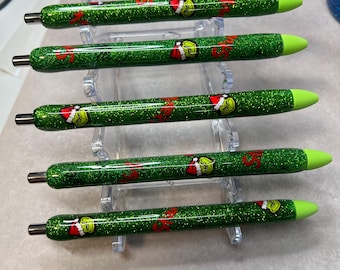 Può includere: Sei penne coperte di glitter a tema natalizio. Le penne sono verdi con un design del personaggio del Grinch e dettagli rossi. Le punte delle penne sono verde chiaro. Le penne sono esposte su un supporto di plastica trasparente.
