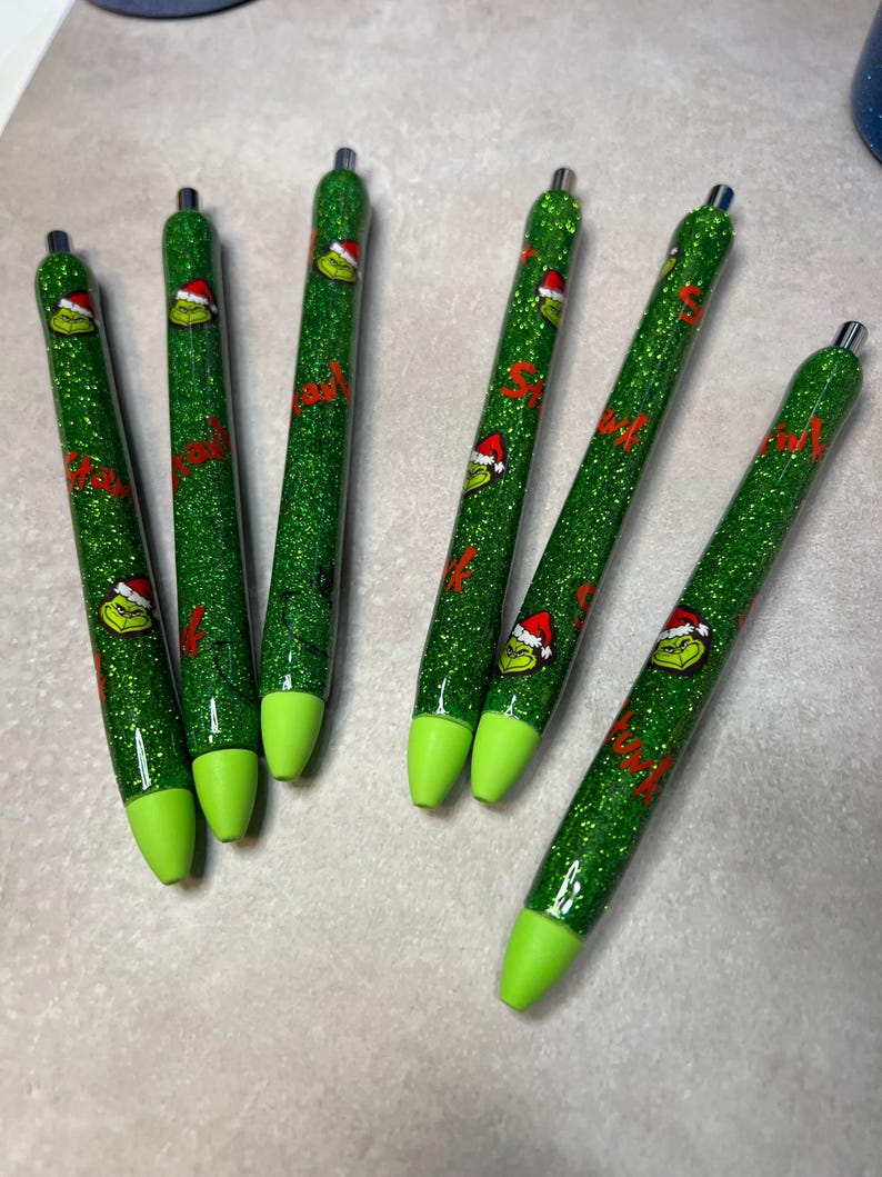 May include: Six green glitter pens with a Grinch design. The pens have a green glitter barrel, a lime green tip, and a silver metal top. The Grinch is wearing a Santa hat. The word "Smile" is written in red on the pens.