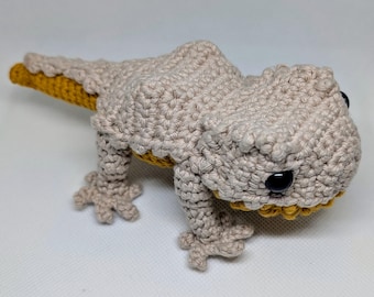 May include: A crocheted beige and yellow lizard toy with black eyes. The lizard is standing on a white surface.