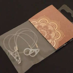 May include: A clear plastic package containing two clear plastic ear hooks and a clear plastic cord. The package is on top of a brown card with a gold mandala design.