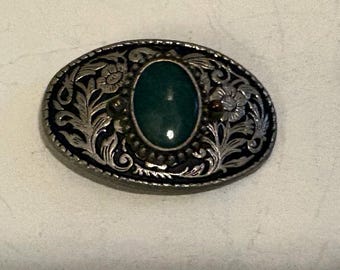 May include: Oval-shaped belt buckle with a dark silver border and a central green stone. The buckle features floral and leaf designs, with a detailed, engraved pattern. The buckle is a vintage style.