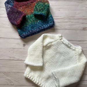 May include: Two knitted baby sweaters, one with a rainbow pattern and one white.