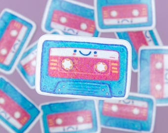 May include: A blue and pink cassette tape sticker with a smiling face. The sticker has a glittery texture and is surrounded by other cassette tape stickers.