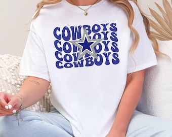 May include: White t-shirt featuring the word "COWBOYS" in blue, stacked three times, with a silver star in the centre. The shirt has a crew neck and short sleeves. The design uses a retro, wavy font.