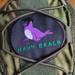 以下が含まれることがあります： 緑色の迷彩柄の戦術バッグに、紫色のアザラシが緑色の目で描かれた黒いパッチが付いており、緑色の文字で「NAVY SEALS」と書かれている。