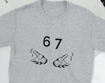 May include: A light gray t-shirt featuring the number "67" above two cartoon hands with curved fingers. The hands are black and appear to be gesturing upwards. The shirt is made of a soft material.