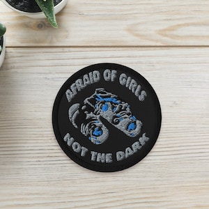 Könnte beinhalten: Ein runder, schwarzer, gestickter Aufnäher mit dem Text "AFRAID OF GIRLS NOT THE DARK" in Weiß und einem blau-grauen Design. Zwei kleine Sukkulenten in Töpfen befinden sich im Hintergrund auf einer hellen Holzoberfläche.