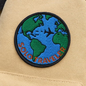 Peut inclure: Sac à dos en toile beige avec un patch brodé représentant un globe terrestre bleu avec des continents verts et un avion blanc. Le patch indique "SOLO TRAVELER" en lettres noires et orange.