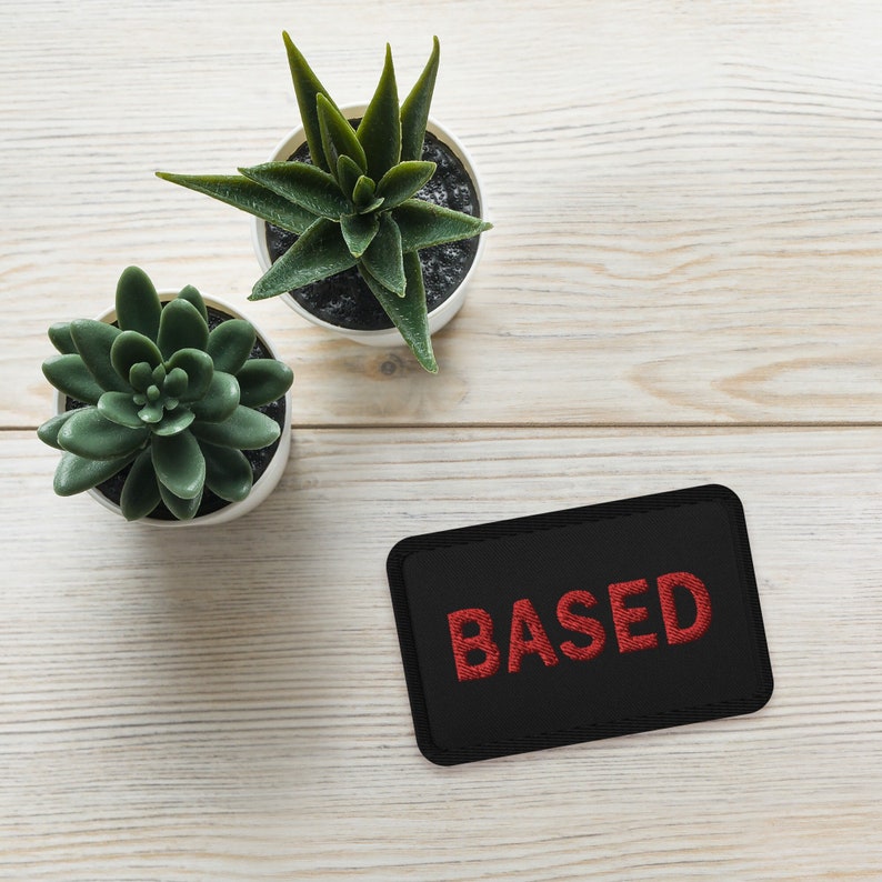Op de afbeelding: Een zwarte patch met het woord "BASED" geborduurd in rood op een wit houten oppervlak met twee potplanten.