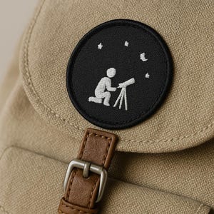 May include: A tan backpack with a black circular patch featuring a white embroidered design of a person looking through a telescope at stars and a crescent moon. The backpack has a brown leather strap and a silver buckle.