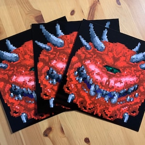 Op de afbeelding: Drie zwarte en rode prints van een gepixelde demon met een enkel oog en scherpe tanden. De demon is rood met een zwarte achtergrond.